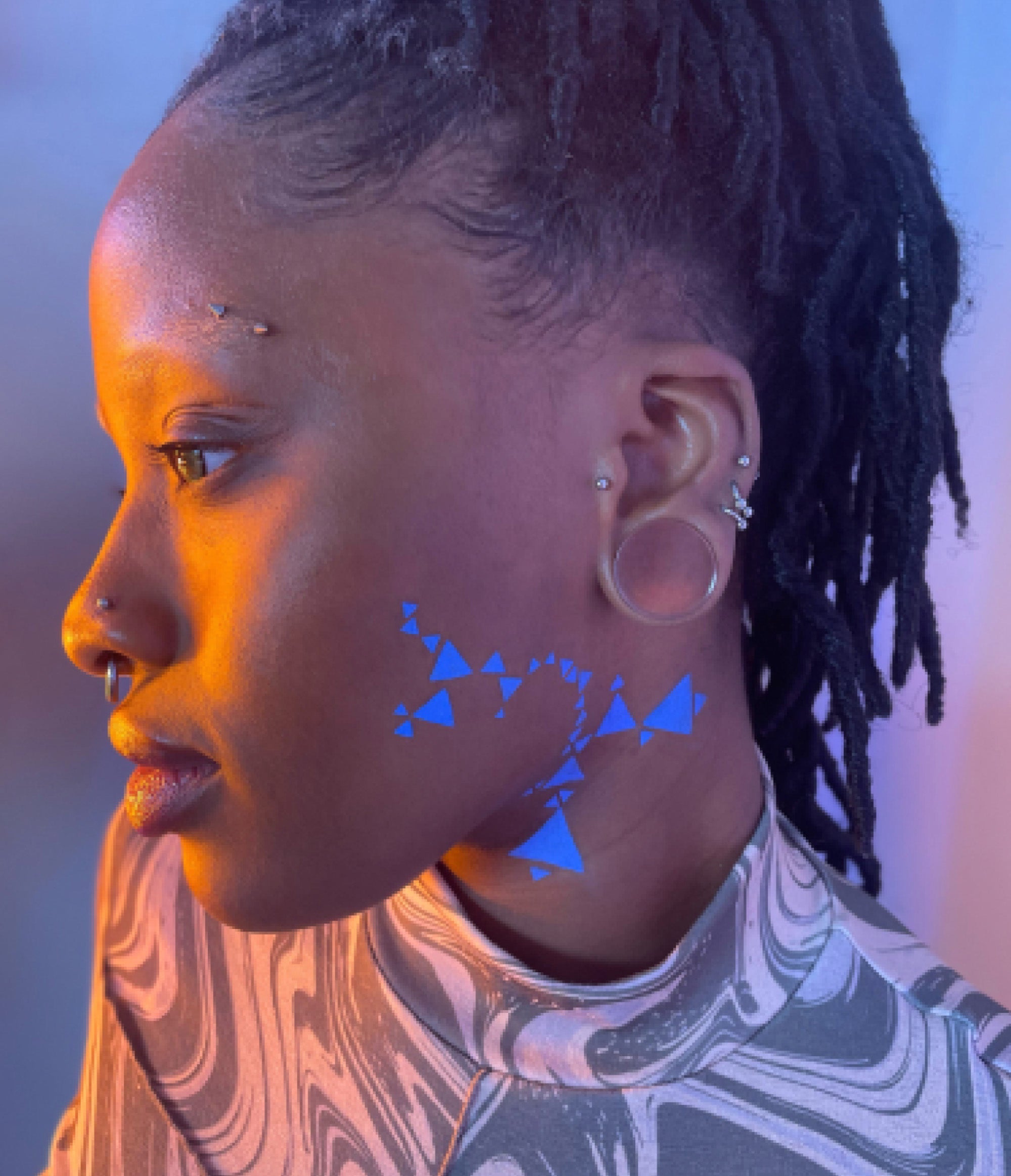 Side view of a model with a dark skin tone wearing Deepdive in Benitoite along their left cheek and neck in a geometeric pattern with triangles.