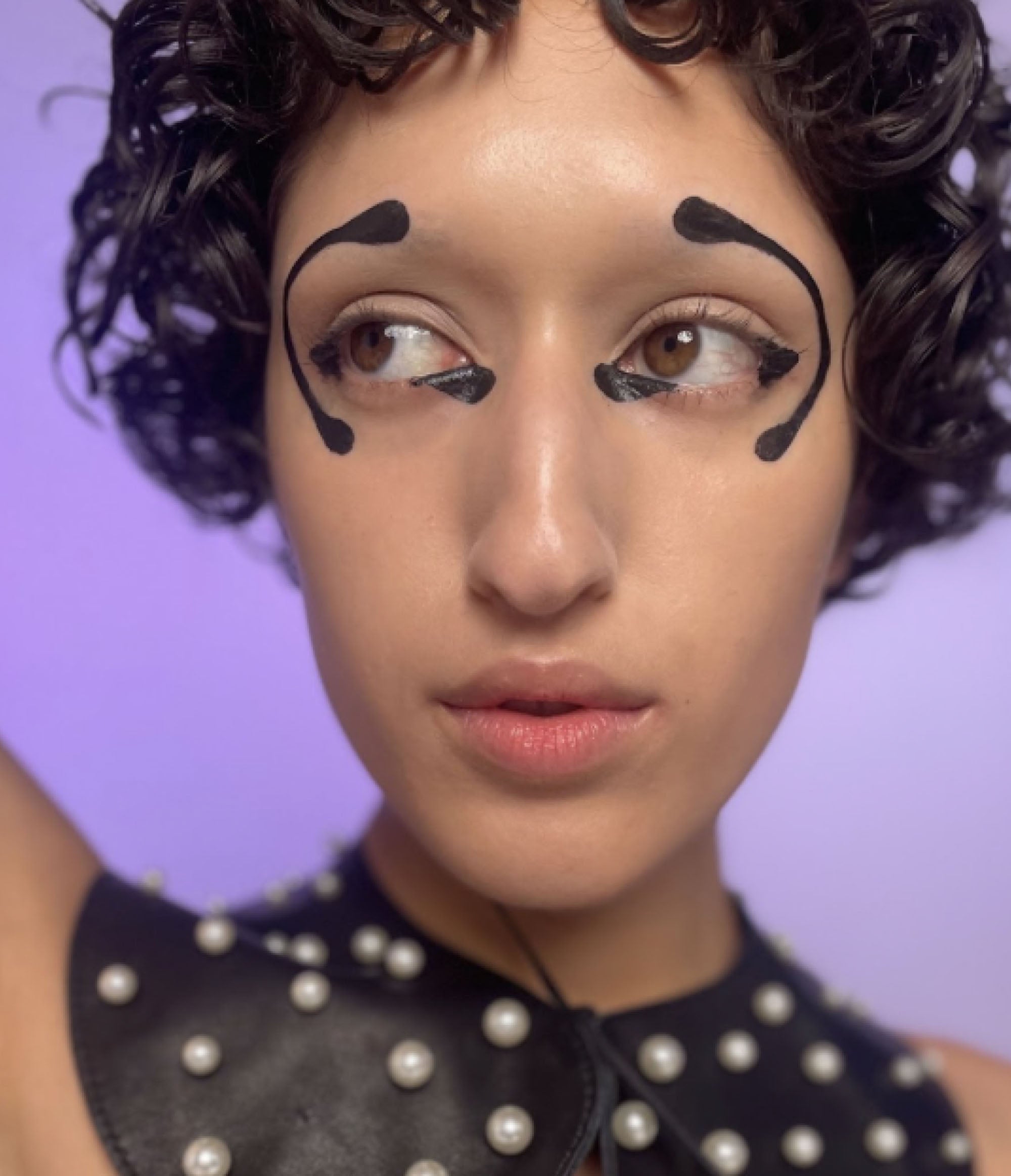 A headshot of a girl with no eyebrows and short brown curly hair wearing a humanoid makeup look. The look consits of a half circle on the outside of each eye that sweeps from the eyebrow to the cheekbone, and black geometric shapes in the corner of each eye