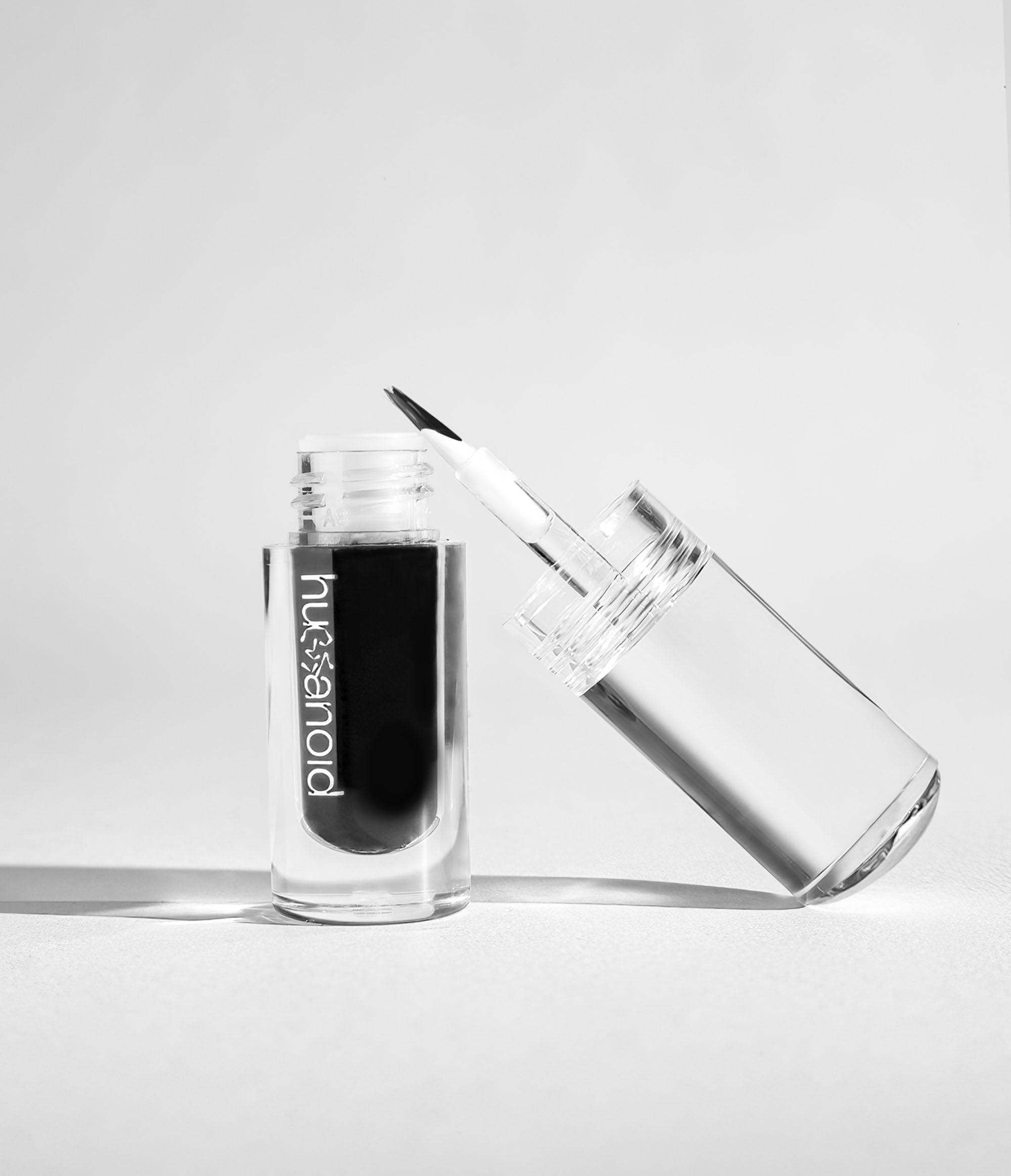 A transparent cylinder with a white humanoid logo on the front filled with black corvid liquid makeup. Leaning against the cylinder is a transparent cap with a brush at the end. The cap has a silver NFC tag on the top. 