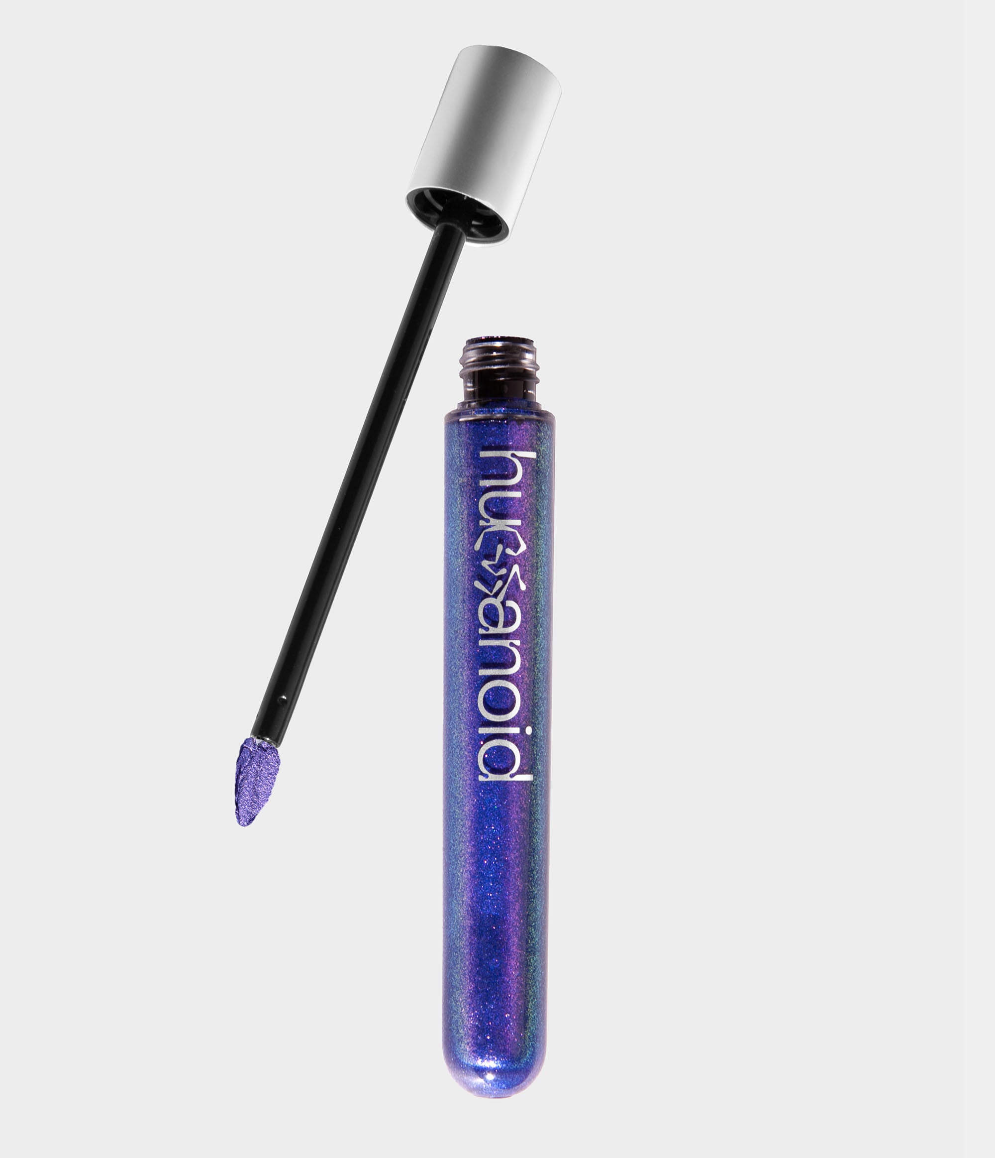 A transparent tube of shimmery vibrant purple liquid makeup with an unscrewed silver cap sitting next to it. A doe foot makeup applicator coated in green makeup extends out of the cap.