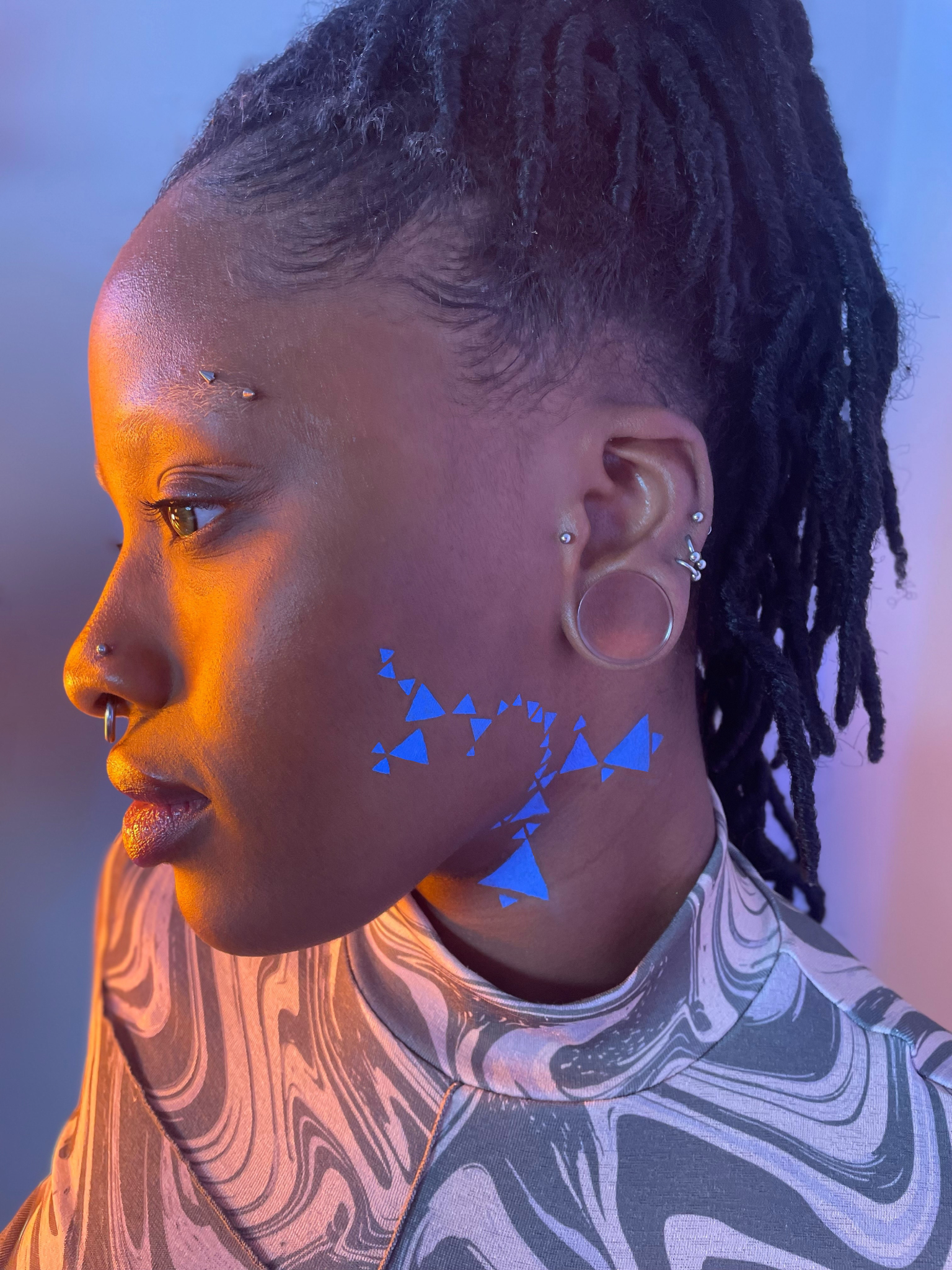 Side view of a model with a dark skin tone wearing deep dive in Benitoite along their left cheek and neck in a geometric pattern with triangles.