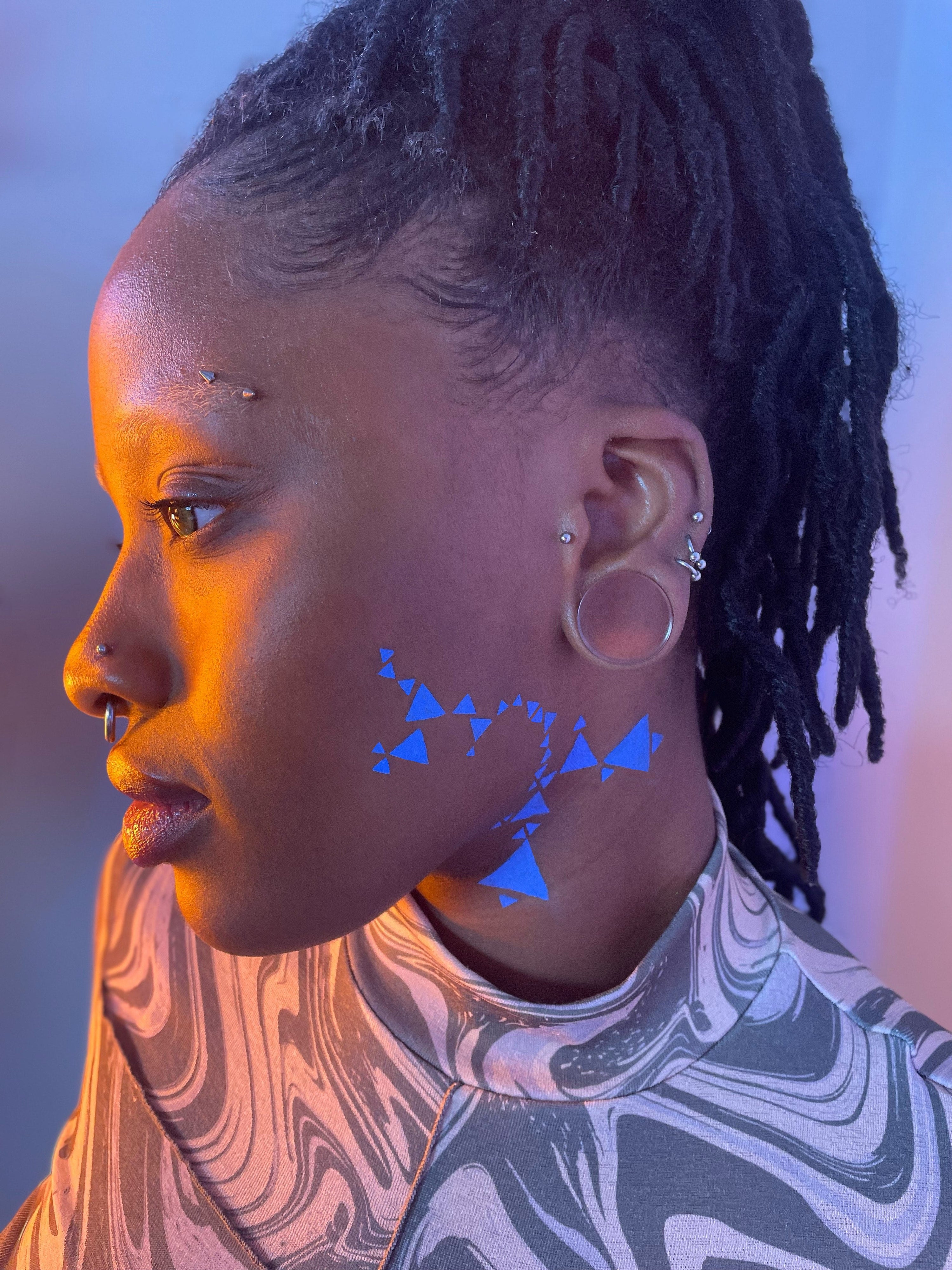 Side view of a model with a dark skin tone wearing deep dive in Benitoite along their left cheek and neck in a geometric pattern with triangles.