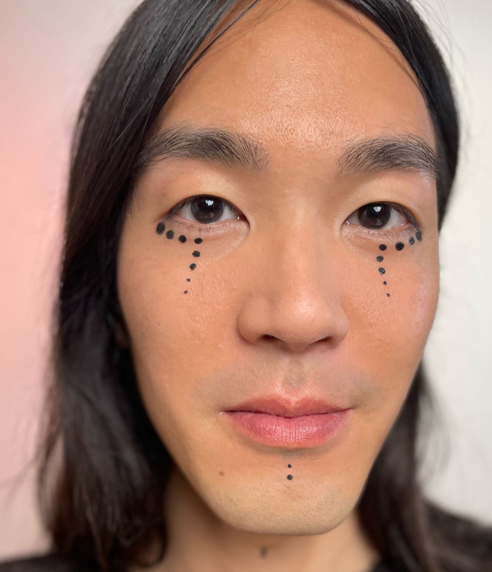 A headshot of a boy with long black hair, and black eyes with a humanoid makeup look on his eyes and chin. The look consits of eight black dots the follow his lower lid line then drop down towards the nose, and two black dots on his chin.